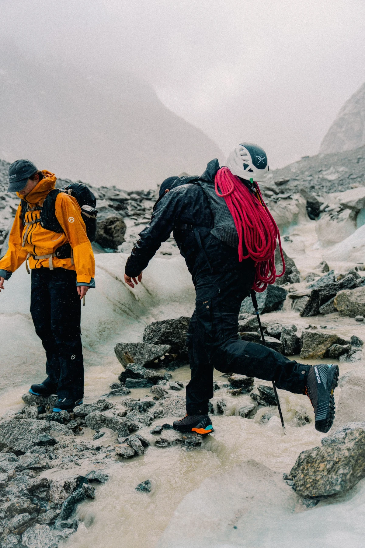 Two climbers crossing wet moraine beside glacial ice, one carrying a coiled rope