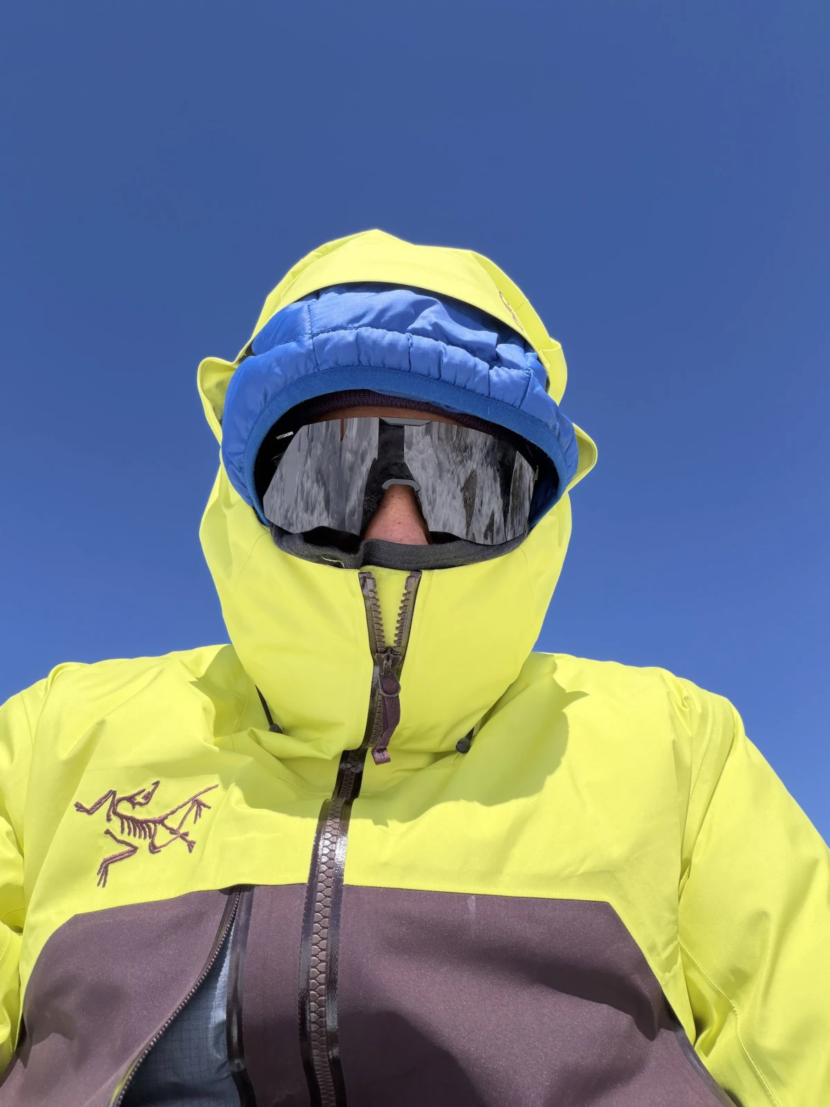 A climber in a bright shell and glacier goggles under a clear blue sky
